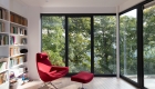 Photograph of the interior view of the writing room, a large red chair is positioned with views out towards the greenery beyond. The room has large full height glazing and sliding doors opening onto raised terrace deck.