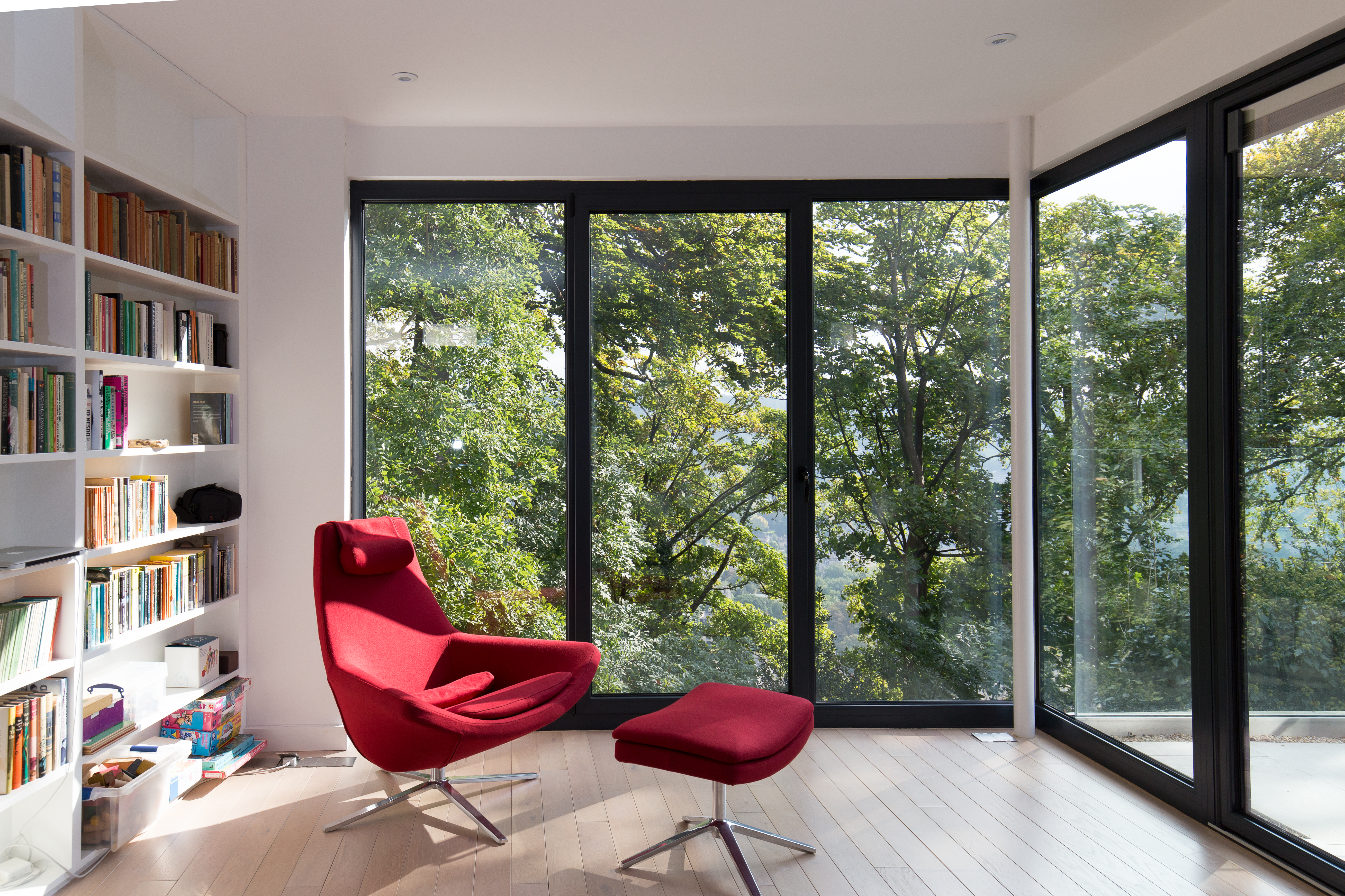 Photograph of the interior view of the writing room, a large red chair is positioned with views out towards the greenery beyond. The room has large full height glazing and sliding doors opening onto raised terrace deck.
